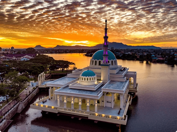 Mosque with turquoise domes and tall minaret stands on water at vibrant sunset.