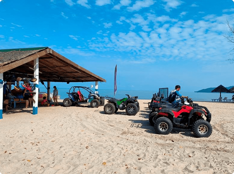 ATVs and dune buggies parked on a sandy beach with people under a wooden shelter.