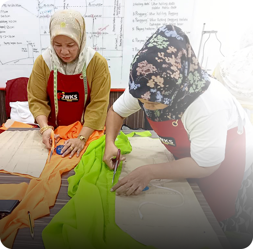 Two women in hijabs cut colorful fabric, focused on sewing work in front of a whiteboard.