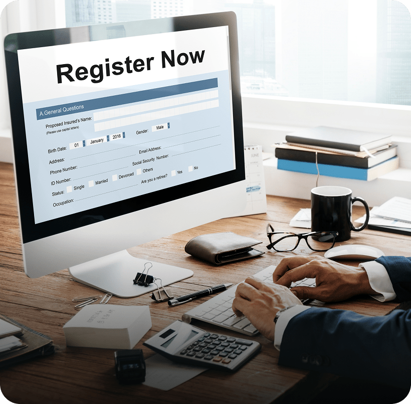 Person in formal wear types a registration form on a computer, surrounded by work essentials.