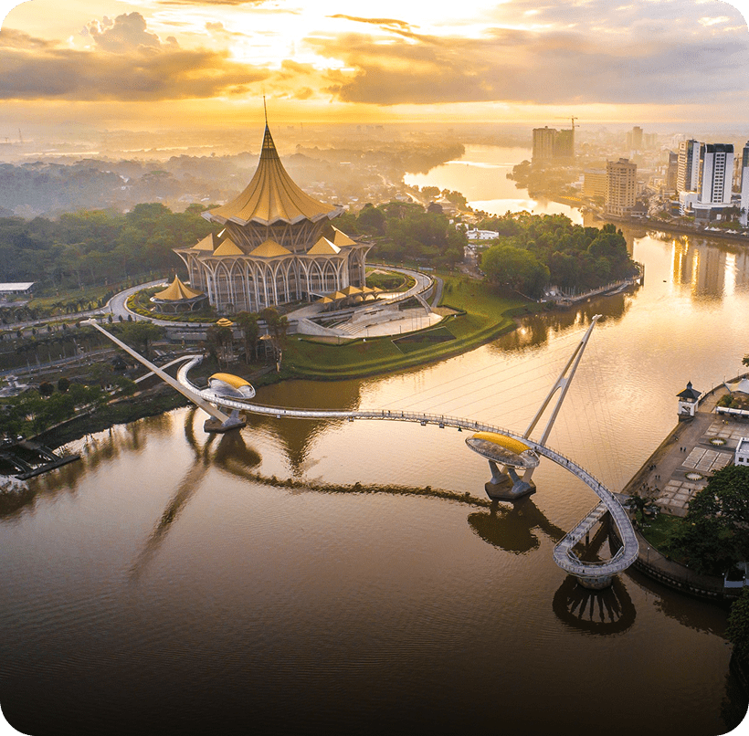 Aerial sunset view of a city with a golden-roof building, modern bridge, and lush greenery.