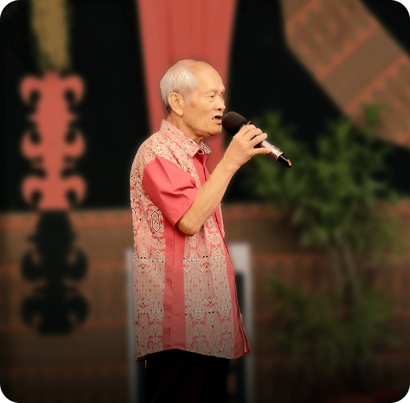 Elderly man in red shirt speaks into a mic at a formal, decorated event.