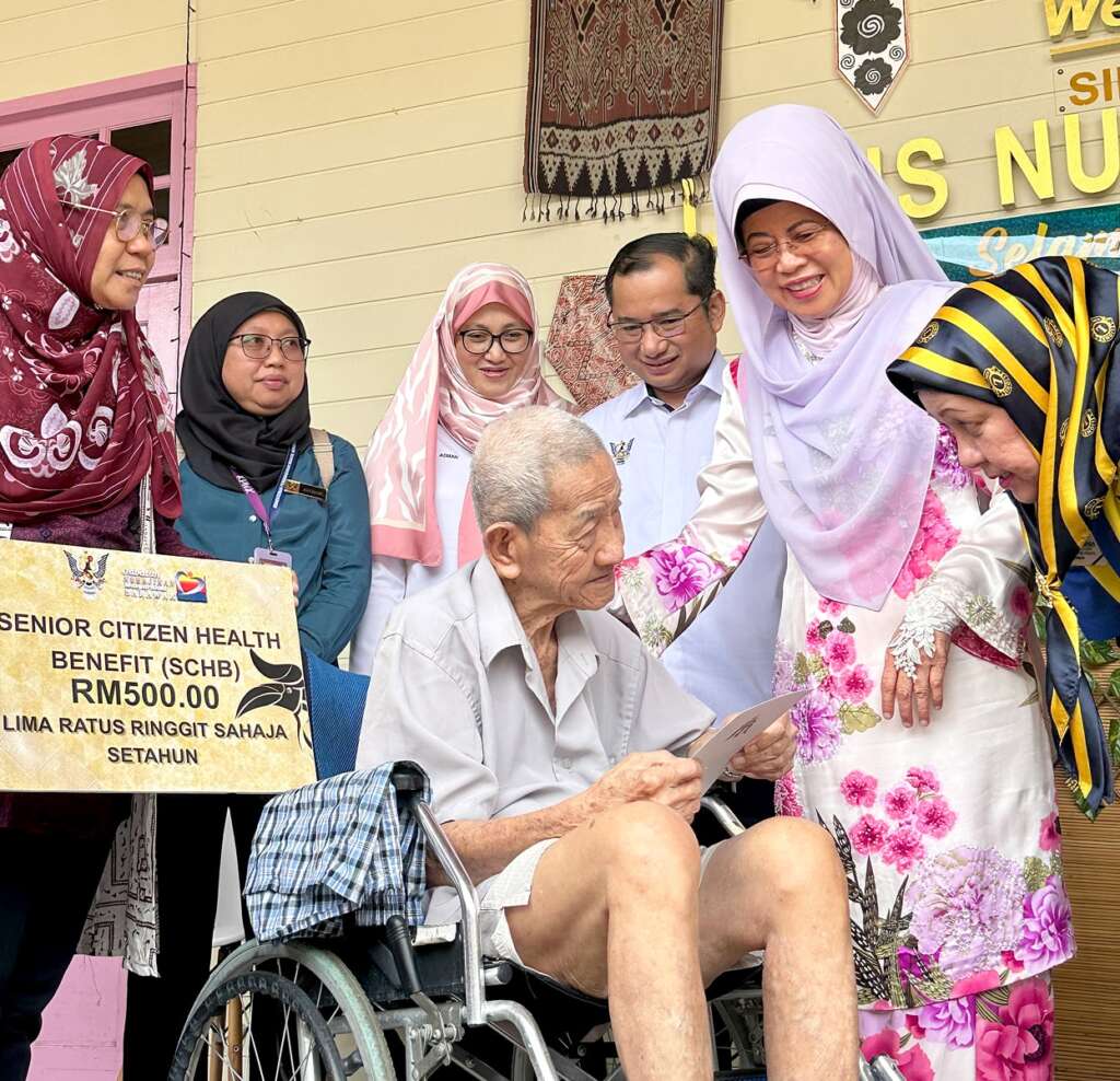 Elderly man in wheelchair receives RM500 health aid, surrounded by smiling supporters.