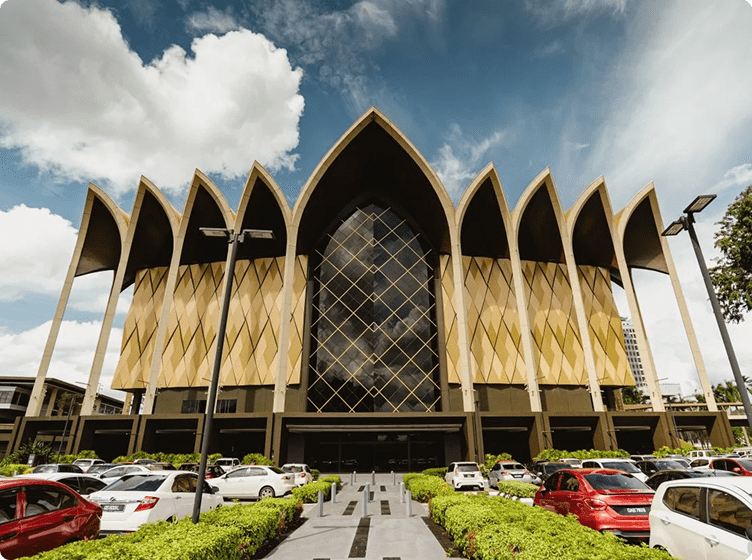 Modern building with pointed arches and diamond windows, cars parked below under a blue sky.