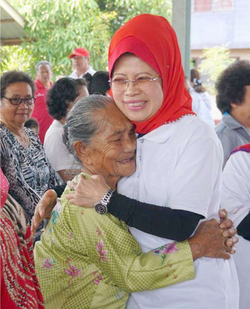 Woman in red headscarf embraces smiling elderly woman in green blouse at outdoor gathering.